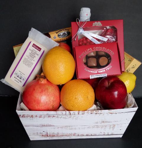 Fruits and chocolates gift basket arranged in a white rustic wooden box on a black background. Oranges, red apples, cheese, a bottle of sparkling cider, and chocolates.