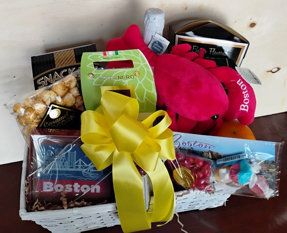 A Boston-themed gift basket filled with snacks, treats. This white wicker basket is decorated with a large yellow satin bow and contains items such as a bag of gourmet caramel popcorn, local handmade chocolate, a red Boston plush toy, and packaged candy and chocolates.