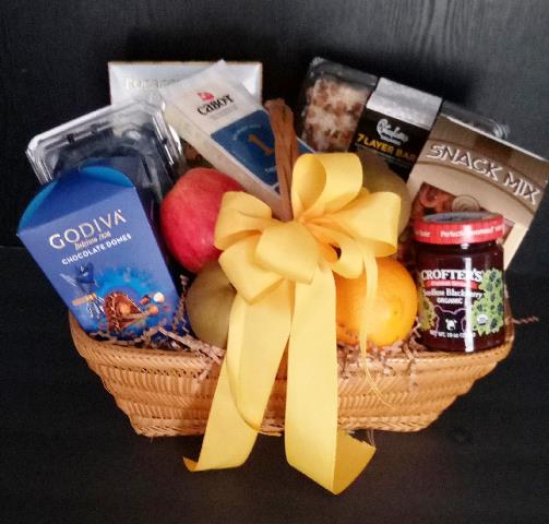 A wicker gift basket filled with assorted snacks and treats, tied with a large yellow ribbon. Items include chocolate gift boxes, chocolate bars, a bag of Snack Mix, fruit jam, and fruits. 